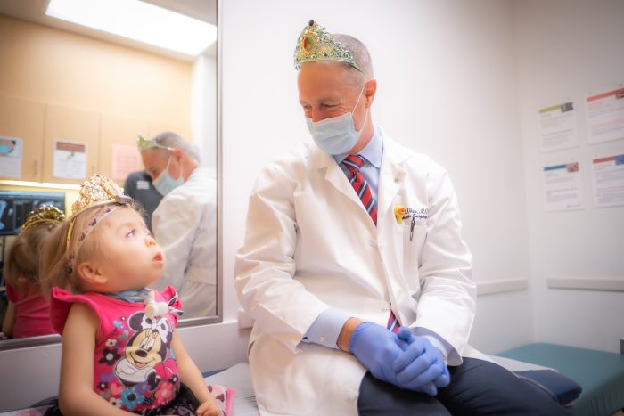 A man wearing a doctor's white coat and a tiara smiling and looking at a toddler, who is looking back at the man.