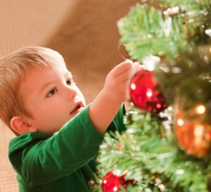 Young blond boy wearing a green shirt places a red glass ball shaped ornament on a Christmas tree