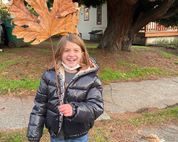 A girl with a wide smile wearing a thick winter jacket and holding a large leaf.