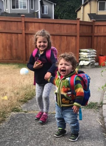 A boy in the foreground and a girl both with wide smiles standing in a yard and wearing backpacks.