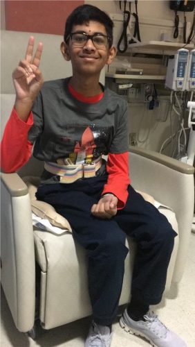 A child smiling and wearing glasses sitting in a medical office giving the peace sign.