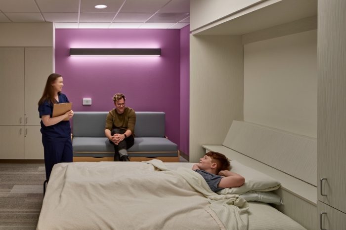 A child in a bed with a medical professional standing to the left and another person sitting on a couch in the background.