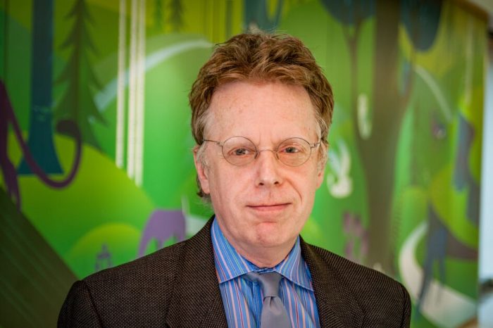 A man wearing glasses and a jacket and tie in front of a mural with green colors.