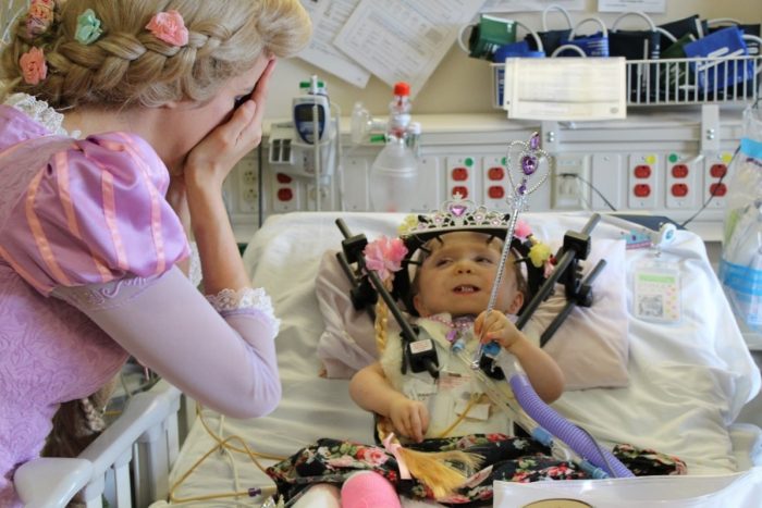 A toddler lying on a medical bed with a device on her head, and a woman on the right dressed as a princess.
